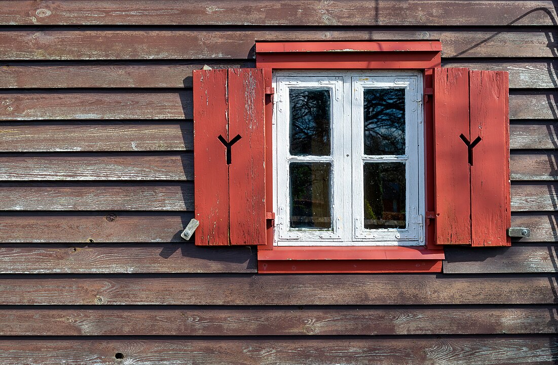 Photo d’une fenêtre à l'encadrement blanc, aux volets rouges ouverts sur un mur en bois