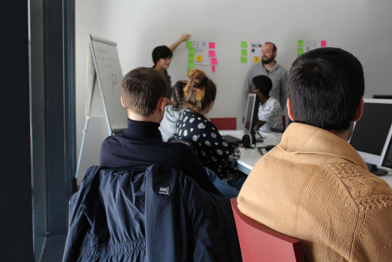 On voit des personnes assises de dos, avec plus au fond Claire qui présente quelque chose avec des post-its colorés sur un mur