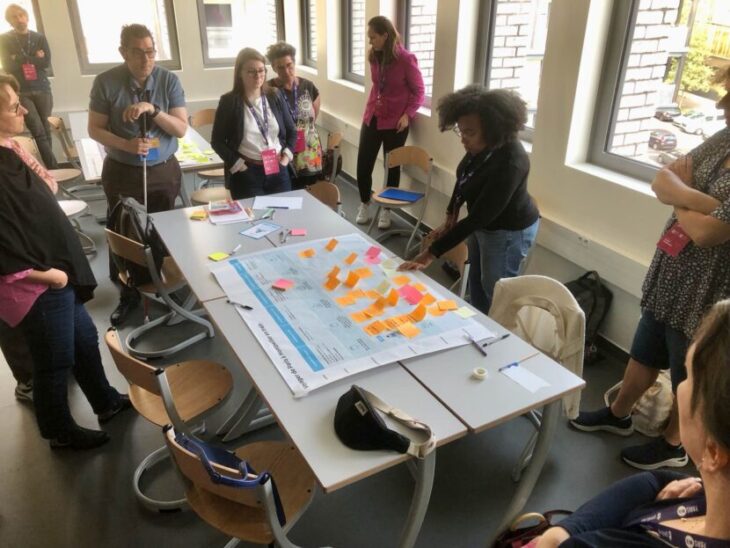 Un groupe de personnes debout autour d'une table sur laquelle sont disposés une grande feuille avec des post-its, crayons et feuilles de papier.